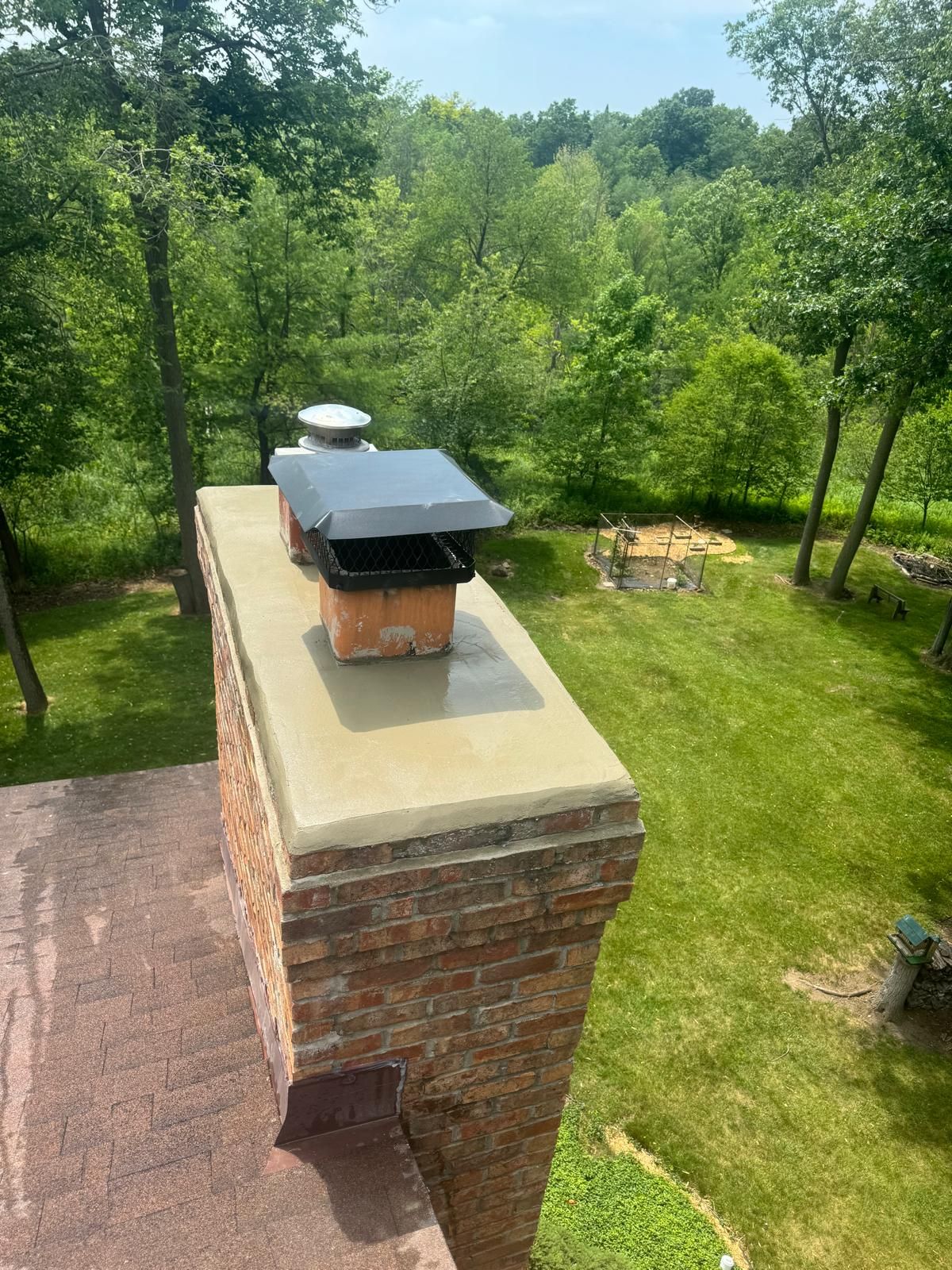 Tall red brick chimney with a smooth mortar crown and a black mesh cap above a tree-lined yard