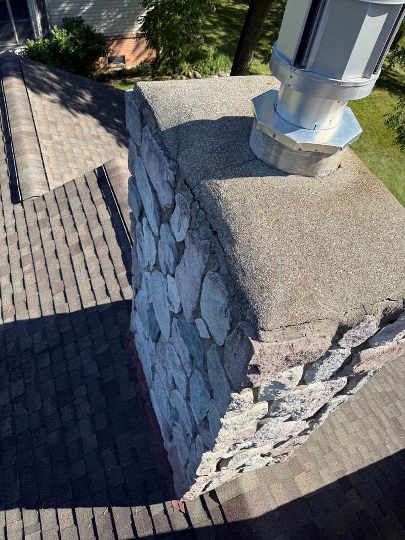 Fieldstone chimney with a cracked, weathered concrete crown and a tall stainless steel vent pipe